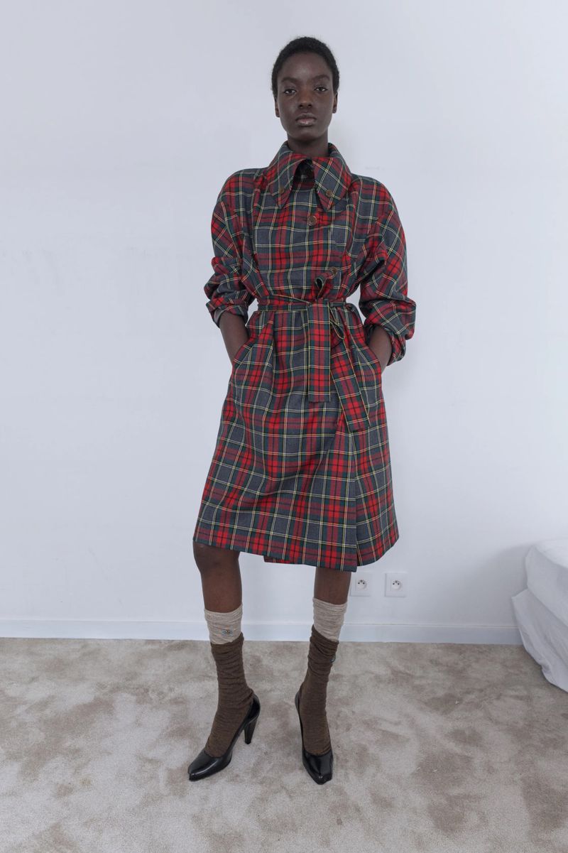 A black female fashion model poses indoors wearing a red, green, and grey plaid dress with brown socks and black high heel shoes.