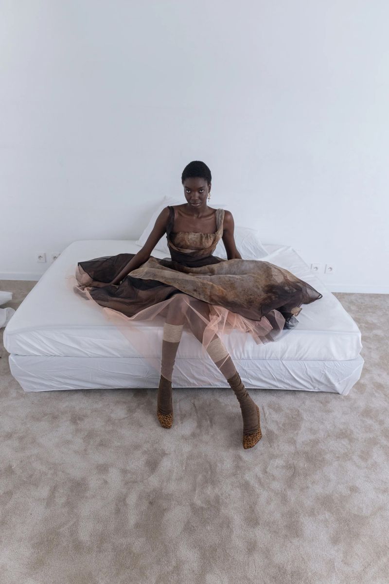 A black female model sits on a white bed in a brown dress, stockings, and heels against a white wall and gray carpet in a studio.