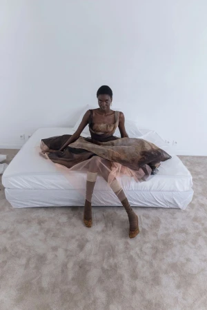 A black female model sits on a white bed in a brown dress, stockings, and heels against a white wall and gray carpet in a studio.