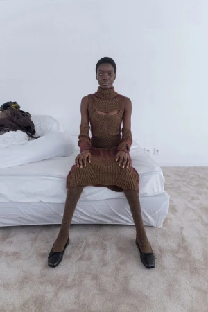 Model seated on a white bed wearing a brown patterned turtleneck dress and matching stockings