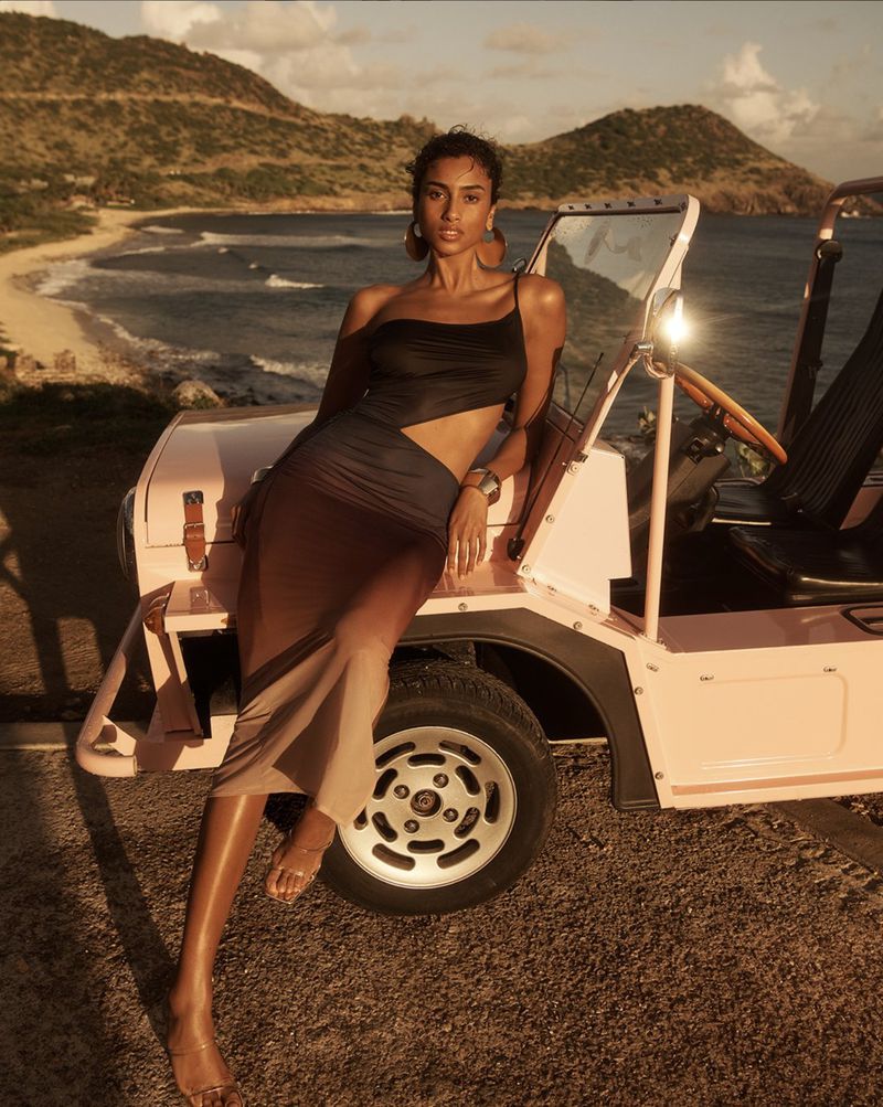 A woman in a stylish cutout dress poses on a pink jeep against a coastal backdrop, showcasing fashion, automotive, and travel themes.