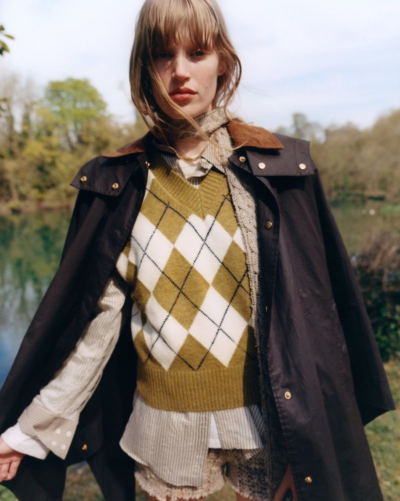 A female fashion model poses outdoors wearing a brown jacket, argyle sweater, and collared shirt in a medium shot on a sunny day.