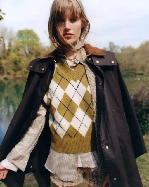 A female fashion model poses outdoors wearing a brown jacket, argyle sweater, and collared shirt in a medium shot on a sunny day.