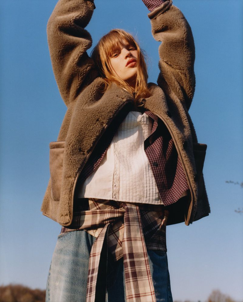 Adult woman with long brown hair poses in sunlight wearing a brown fleece jacket, jeans, and a plaid shirt tied around her waist.