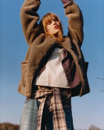 Adult woman with long brown hair poses in sunlight wearing a brown fleece jacket, jeans, and a plaid shirt tied around her waist.