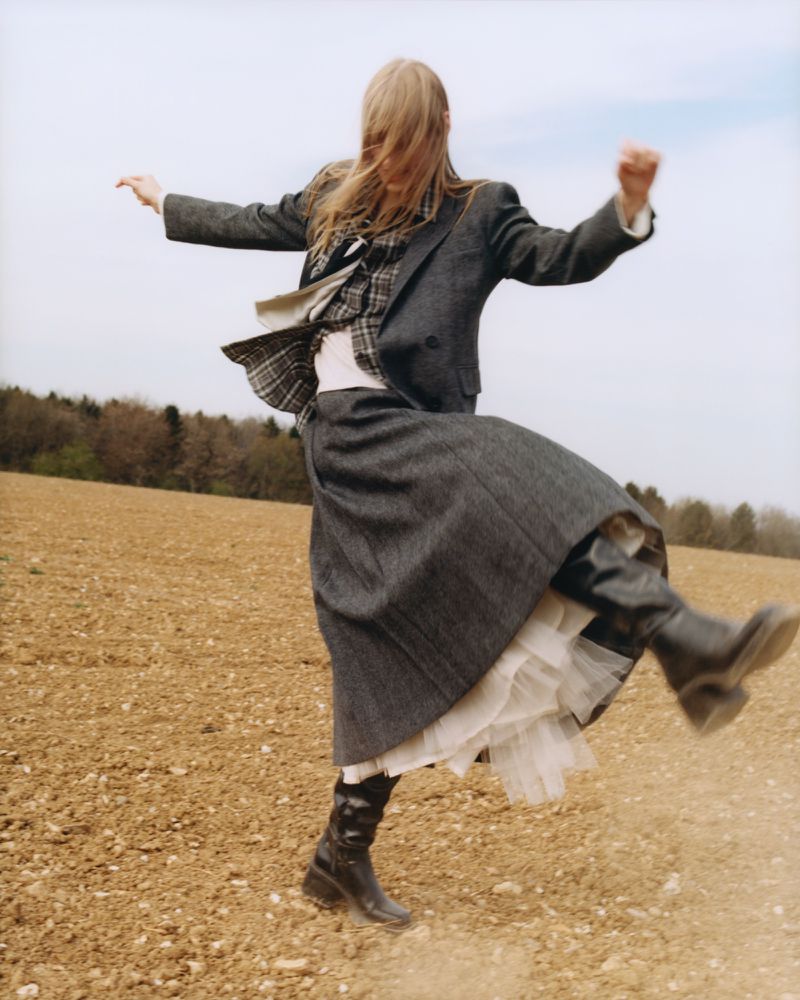 A woman with blonde hair leaps energetically in a field, wearing a grey jacket, skirt, and black boots, against a backdrop of a cloudy sky.