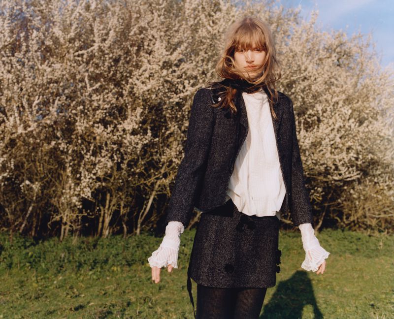 A young woman poses outdoors wearing a tweed skirt suit, tights and ruffled sleeves, set against a blossoming tree and green grass.