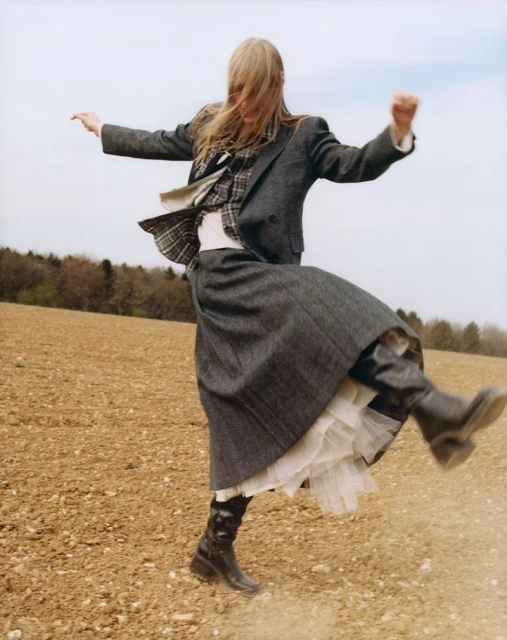 Fashion shot of a woman in a grey blazer and skirt jumping in a field with a sparse tree line under a pale sky, editorial-style image.