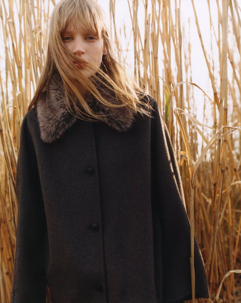A woman with long hair, wearing a dark-colored coat with a fur collar, poses in front of tall, golden reeds in an outdoor setting.