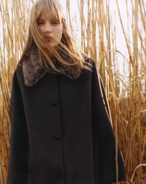 A woman with long hair, wearing a dark-colored coat with a fur collar, poses in front of tall, golden reeds in an outdoor setting.