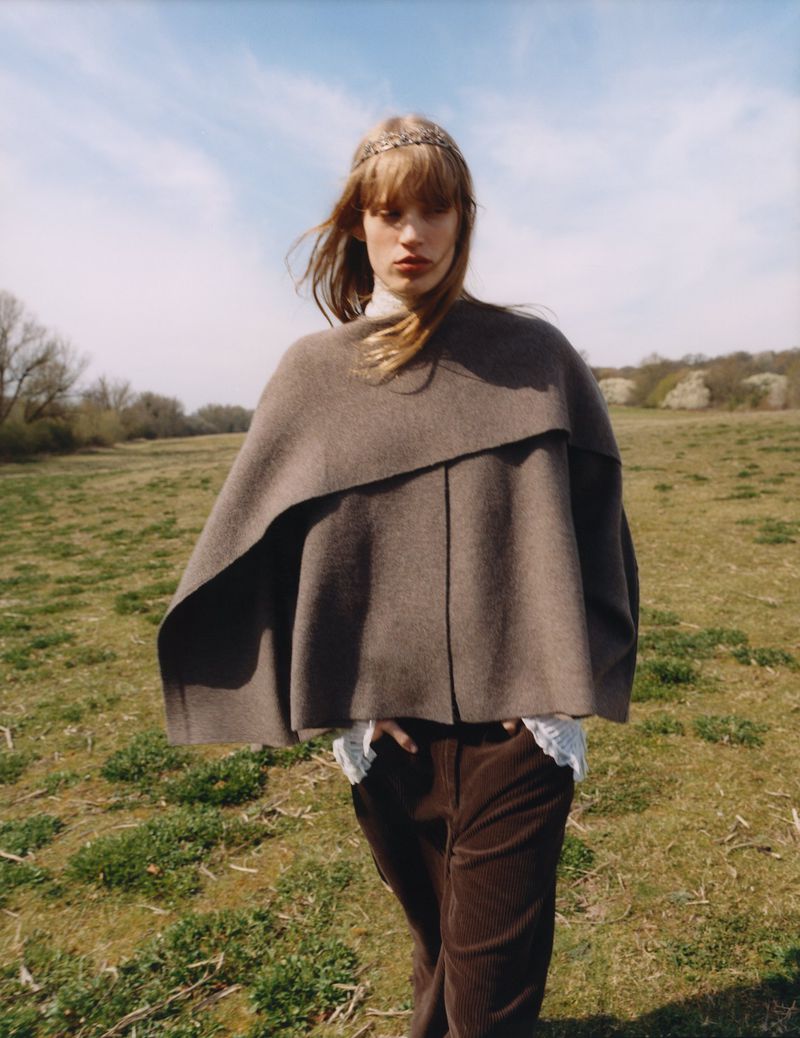 Model in brown poncho and corduroy trousers standing in a grassy meadow under an open sky for Zara April 2025.
