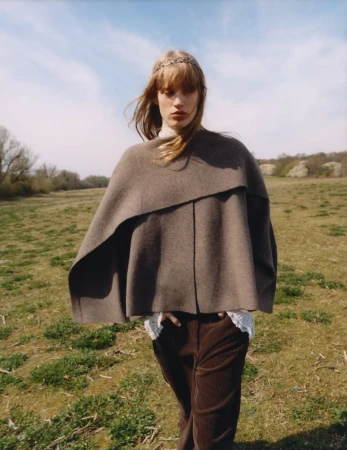 Model in brown poncho and corduroy trousers standing in a grassy meadow under an open sky for Zara April 2025.