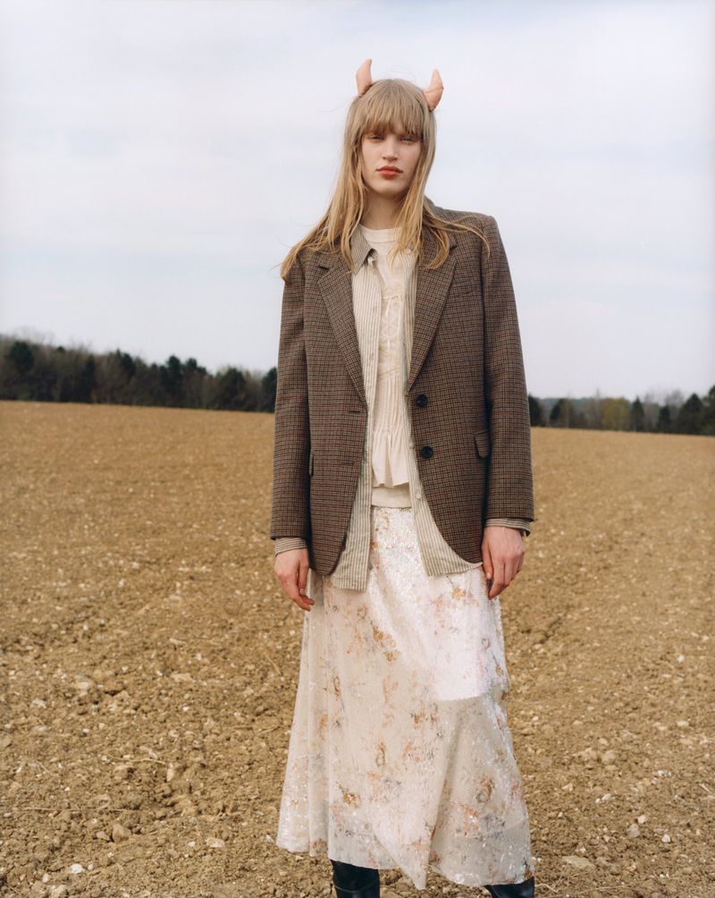 Blonde woman with horn accessory in a field, wearing a tweed blazer, and a long floral skirt. Rural fashion shot.