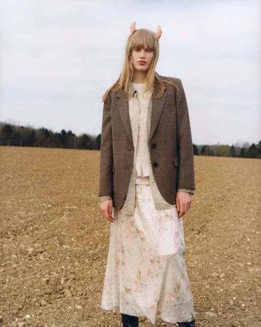 Blonde woman with horn accessory in a field, wearing a tweed blazer, and a long floral skirt. Rural fashion shot.