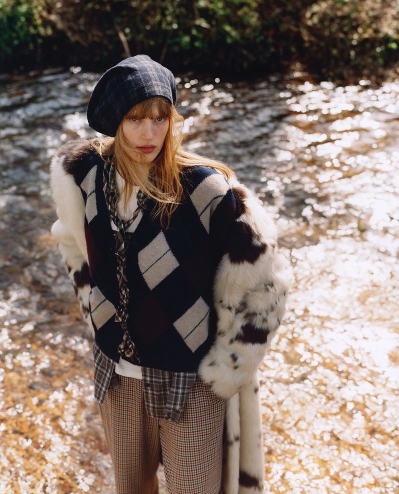 A woman with auburn hair poses next to a stream. She wears a navy argyle sweater, plaid beret, fur-trimmed coat, and patterned pants.