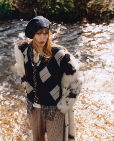 A woman with auburn hair poses next to a stream. She wears a navy argyle sweater, plaid beret, fur-trimmed coat, and patterned pants.