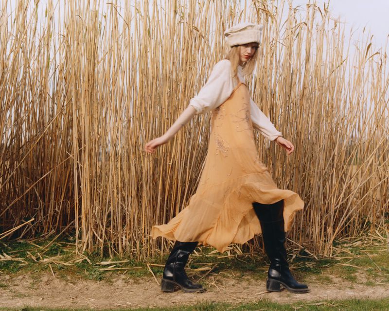 A young woman walks across a grassy field with tall reeds, wearing a long flowing dress, hat, and boots in an outdoor fashion shot.
