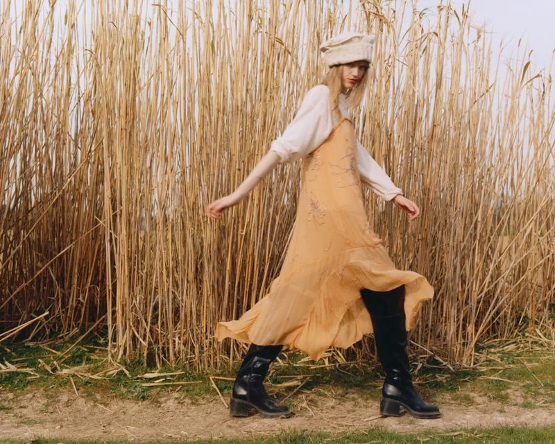 A young woman walks across a grassy field with tall reeds, wearing a long flowing dress, hat, and boots in an outdoor fashion shot.