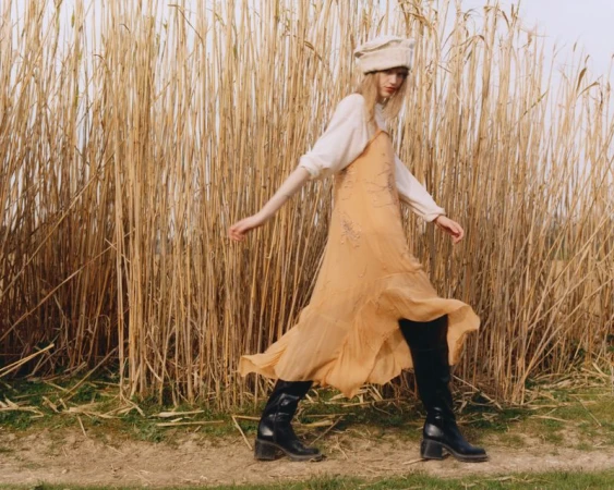 A young woman walks across a grassy field with tall reeds, wearing a long flowing dress, hat, and boots in an outdoor fashion shot.
