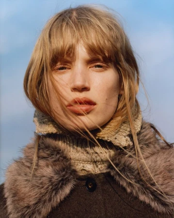 Close-up portrait of a model with blonde hair wearing a dark coat and knitted collar against a clear blue sky