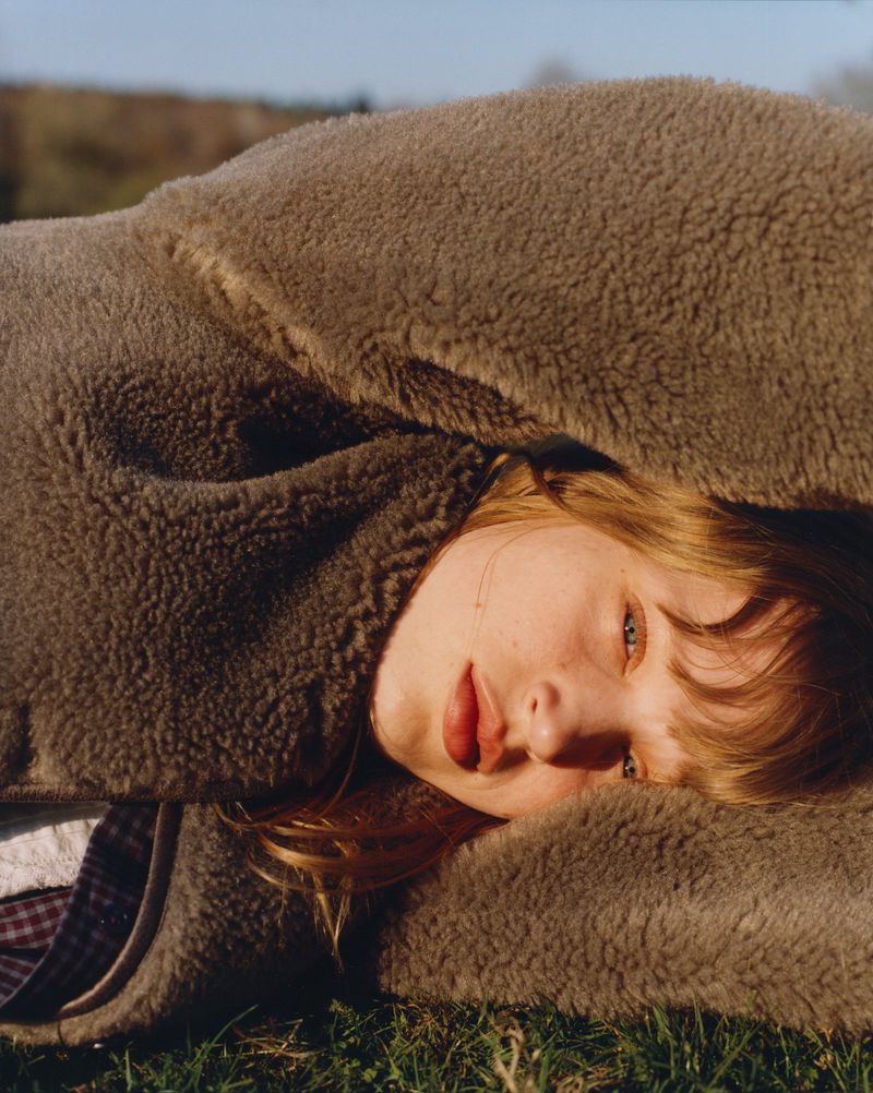 Close-up of a blonde woman lying on grass under a furry brown blanket in natural light, showing her face and a relaxed expression.