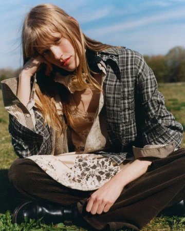 Model with long blonde hair wearing a plaid shirt and brown trousers seated outdoors in a sunny meadow.