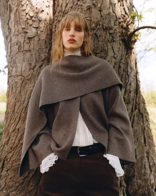 Woman in brown cape poses next to tree in outdoor fashion shot with tiara, corduroy pants, and white lace-trimmed top.