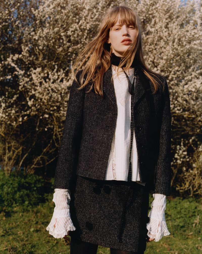 A young woman stands in an outdoor field wearing a dark tweed jacket and skirt with a white blouse and black scarf, set against a blooming tree.