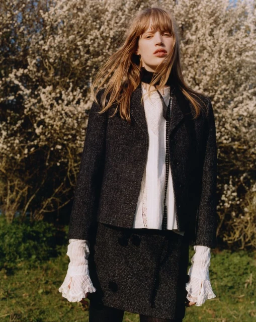 A young woman stands in an outdoor field wearing a dark tweed jacket and skirt with a white blouse and black scarf, set against a blooming tree.