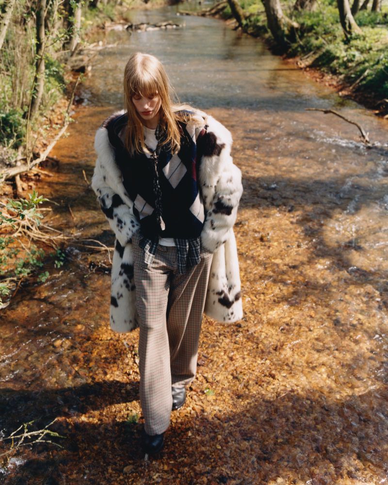 A woman walks in a creek wearing patterned pants, a dark sweater, and a dalmatian-print fur coat, surrounded by lush trees and greenery.