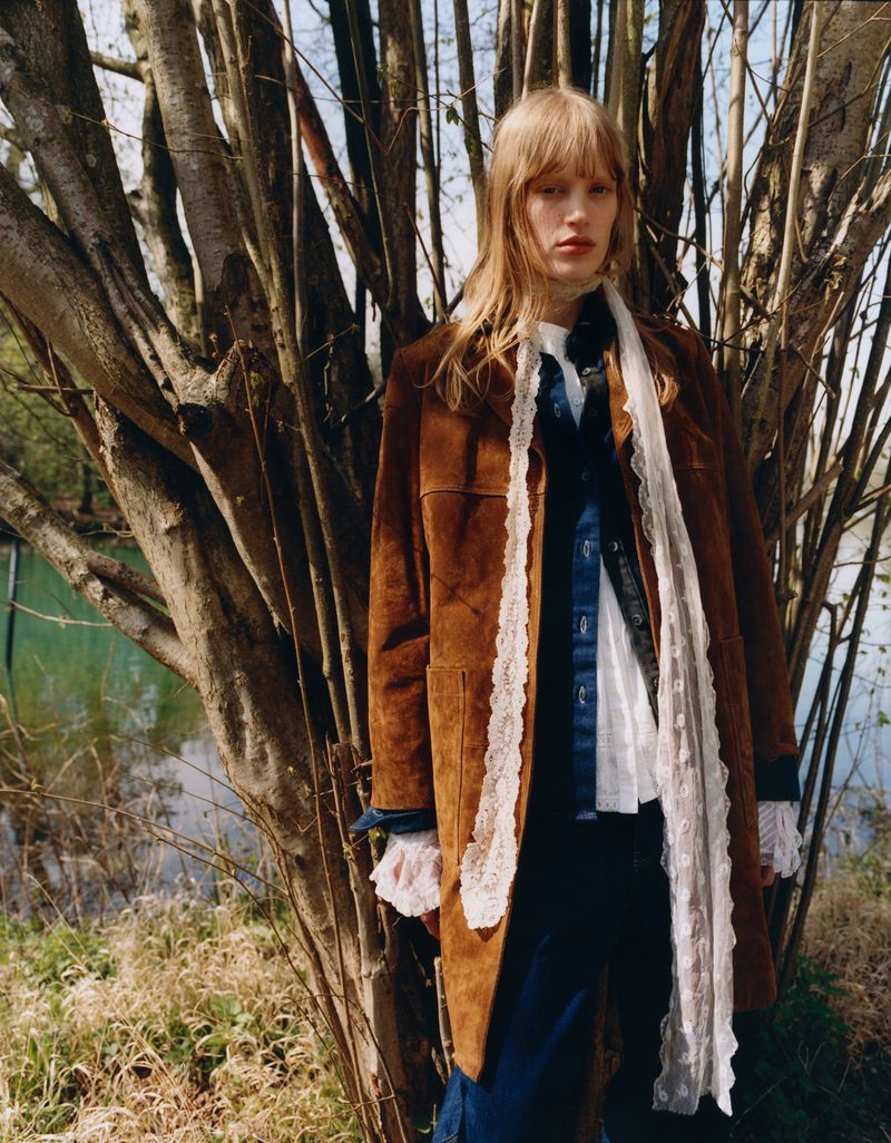 A person with long hair wearing a brown coat and white lace scarf poses outdoors next to a tree with a blurred pond in the background.