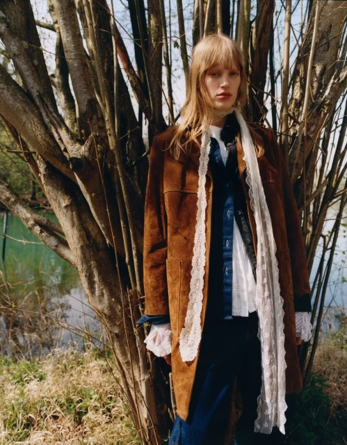 A person with long hair wearing a brown coat and white lace scarf poses outdoors next to a tree with a blurred pond in the background.