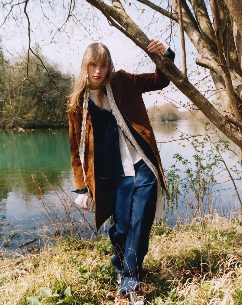 Fashion shot of woman in blue jeans, brown coat, and white blouse, leaning against a tree trunk, with lake and greenery in the background.