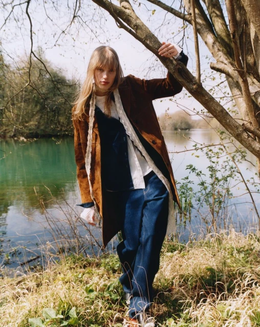 Fashion shot of woman in blue jeans, brown coat, and white blouse, leaning against a tree trunk, with lake and greenery in the background.