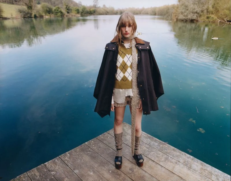 A young female model poses on a wooden dock in a fashion editorial shot wearing earth-tone sweater, black cape, shorts, and leg warmers.