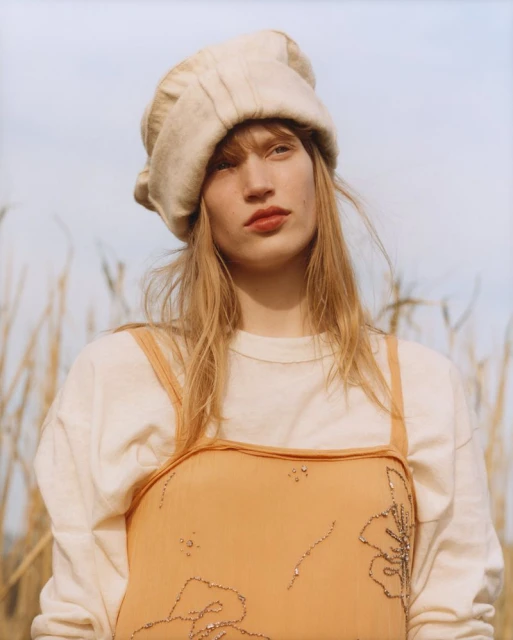 Woman poses in field wearing linen hat, peach dress with floral embellishments, and white long-sleeved shirt in a medium shot.