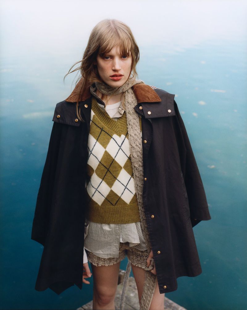 A young woman stands outdoors wearing a dark cape coat, olive argyle sweater, and striped shorts against a blurred water background.