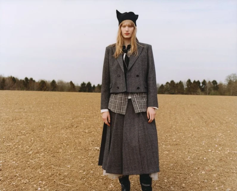 A blonde adult female in a tweed skirt suit and a black hat poses in a field with bare trees in the background on an overcast day.
