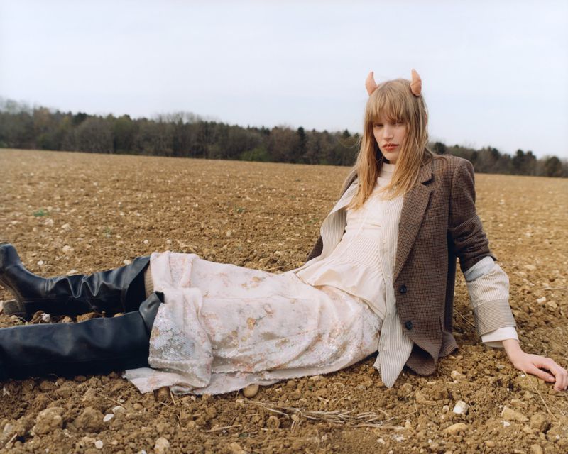 A woman with horns poses in a field wearing a blazer and floral dress. The sky is overcast and a forest is visible in the background.