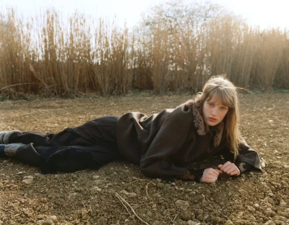 Young woman in brown jacket lies on a brown field, facing the camera with her hands near her face, against a background of tall grass.