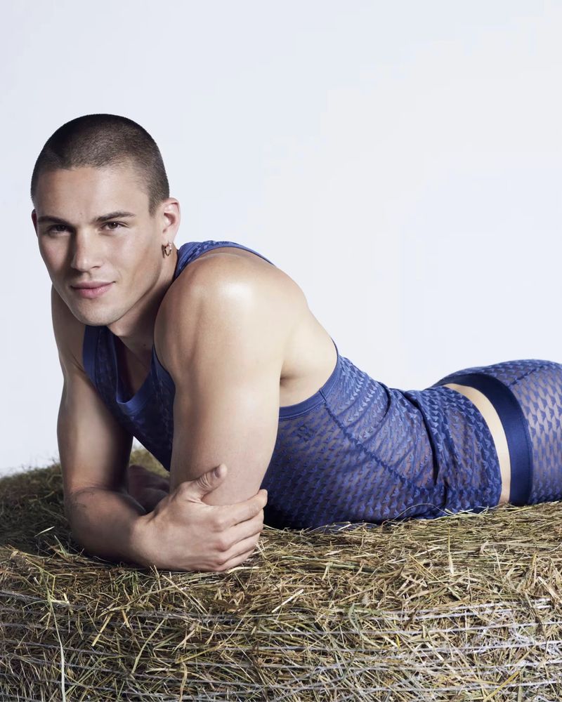 A male model with a buzzcut, wearing a blue mesh top and bottoms, is laying on a bale of hay in a bright, neutral studio.