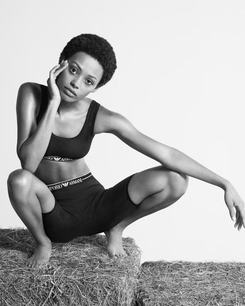 Athletic woman in sports bra and biker shorts poses on hay bales in a studio, showcasing a blend of fashion and fitness in monochrome.