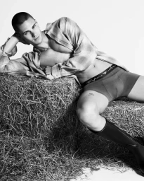 A man wearing underwear and tall boots reclining on stacked bales of hay in a monochromatic studio setting.
