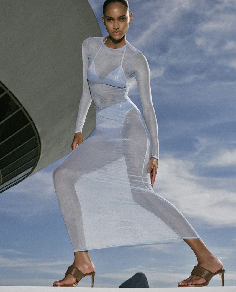 Woman in sheer long-sleeved dress poses on rooftop against a cloudy blue sky near concrete architecture, wearing brown high heels.