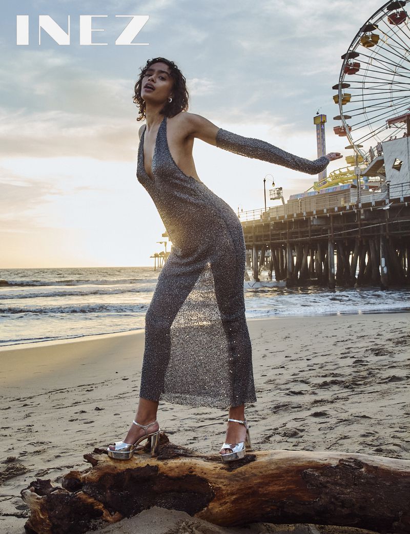 Woman in a glittering dress poses on a log at the beach with a pier and a ferris wheel in the background at sunset.