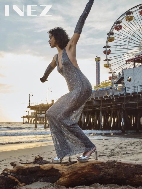 A woman in a sparkling gown poses on the beach with a Ferris wheel and pier in the background at golden hour for a fashion editorial.