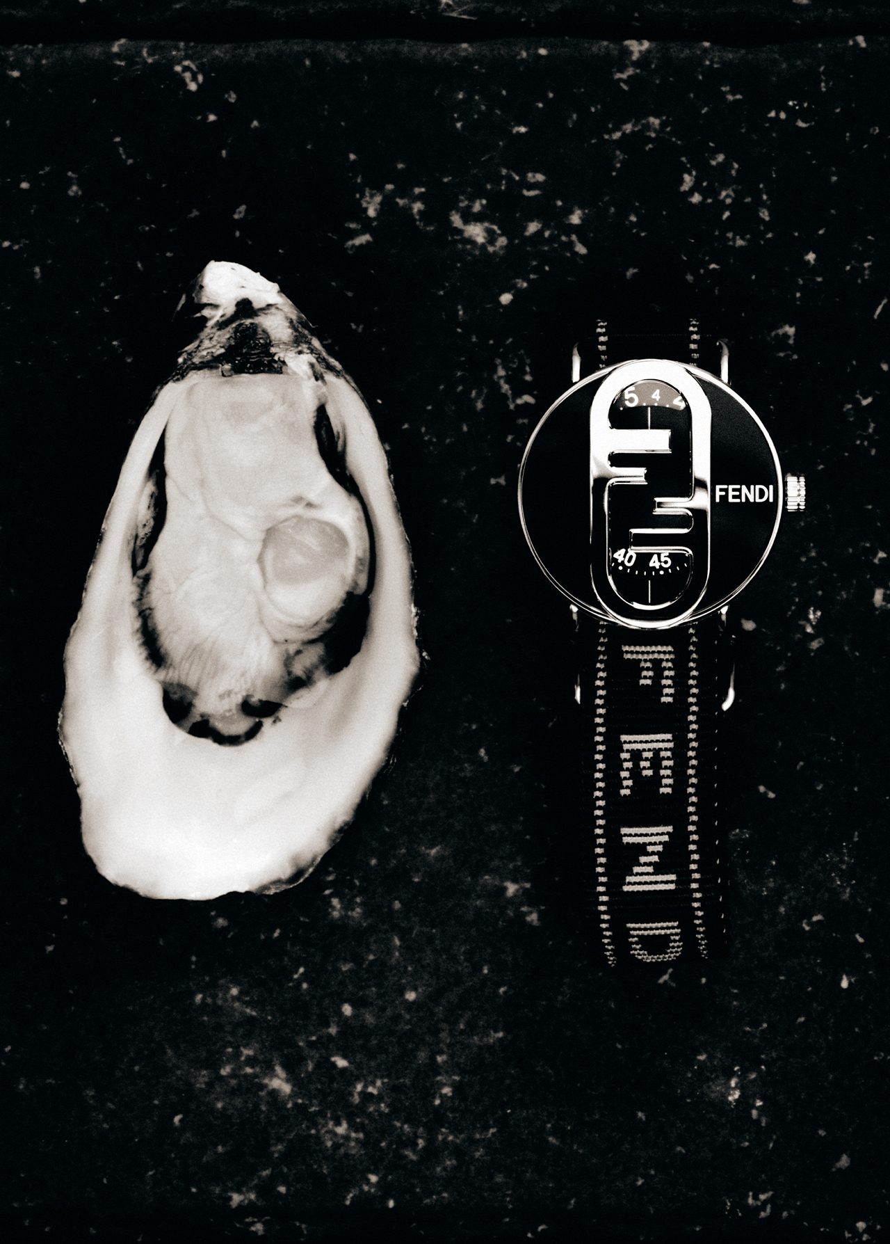 A black and white product shot of an oyster shell and a Fendi watch lying on a mottled black surface, high contrast and moody.