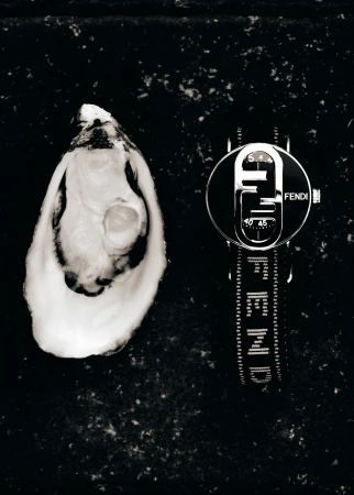 A black and white product shot of an oyster shell and a Fendi watch lying on a mottled black surface, high contrast and moody.