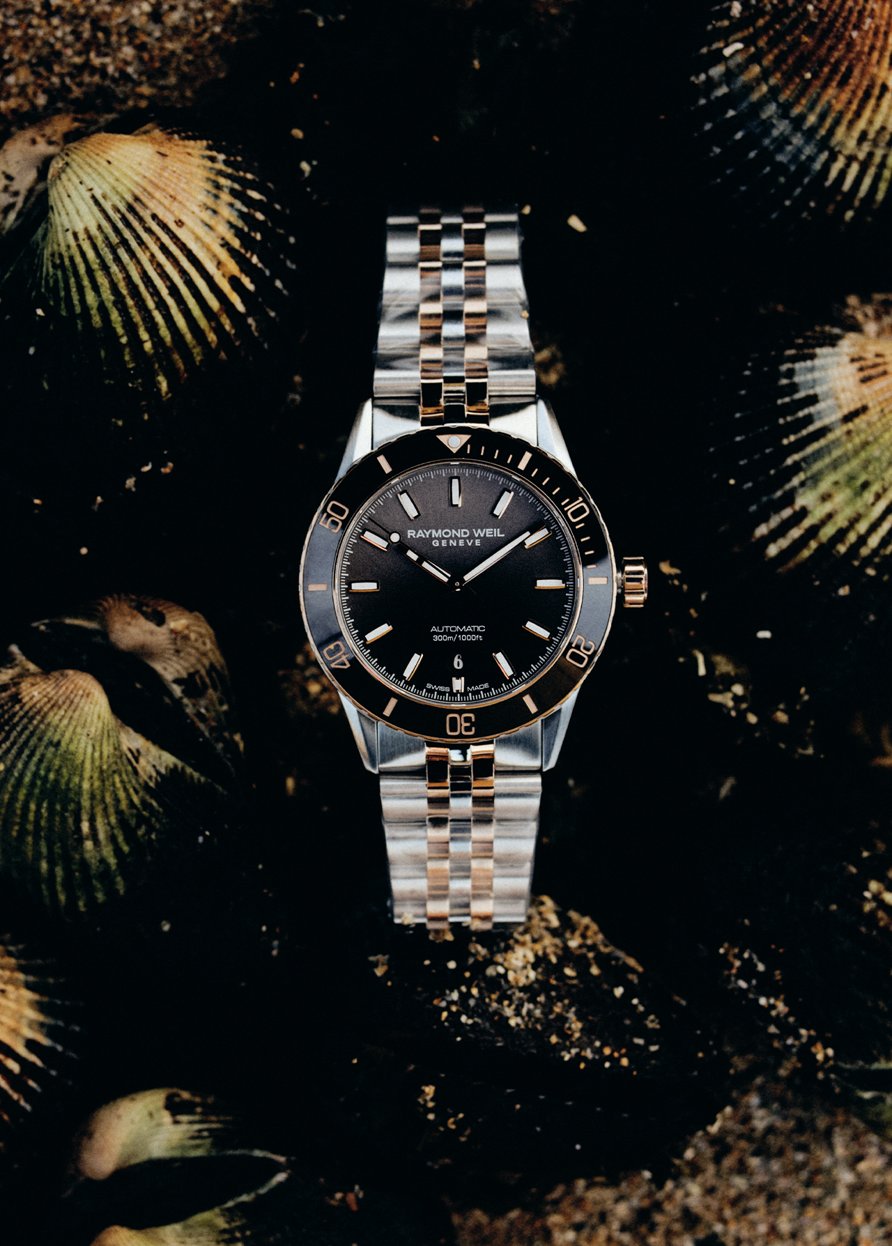 Raymond Weil watch on a two-tone bracelet with a dark face and gold accents displayed on a dark background with seashells.