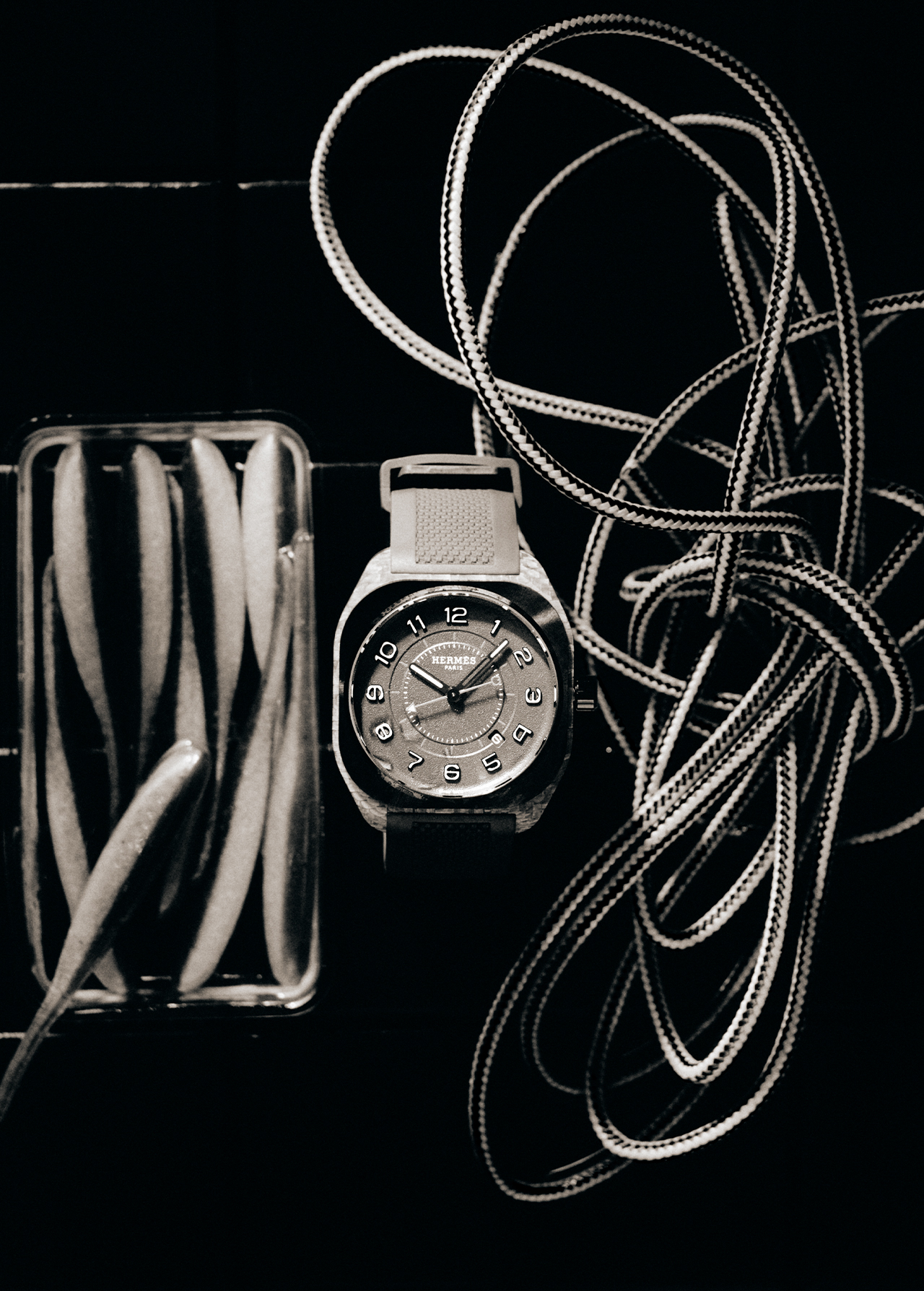 Still life photo of a wristwatch next to a clear container filled with oval shapes, and rope against a dark background.